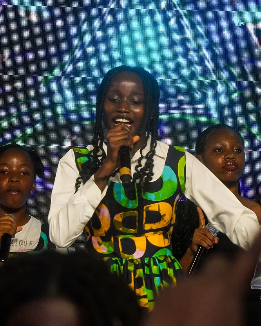 A girl in a green and gold print dress singing on stage at Children’s Day